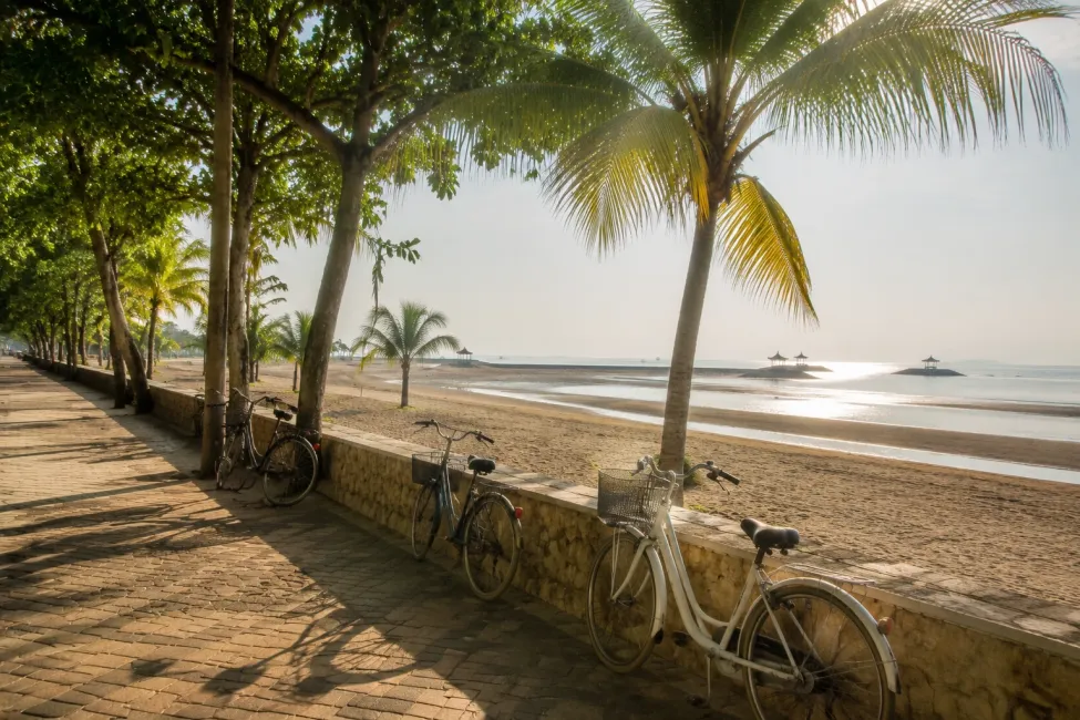 A shaded beachfront promenade in Sanur with bicycles parked along a low wall, palm trees, warm morning light